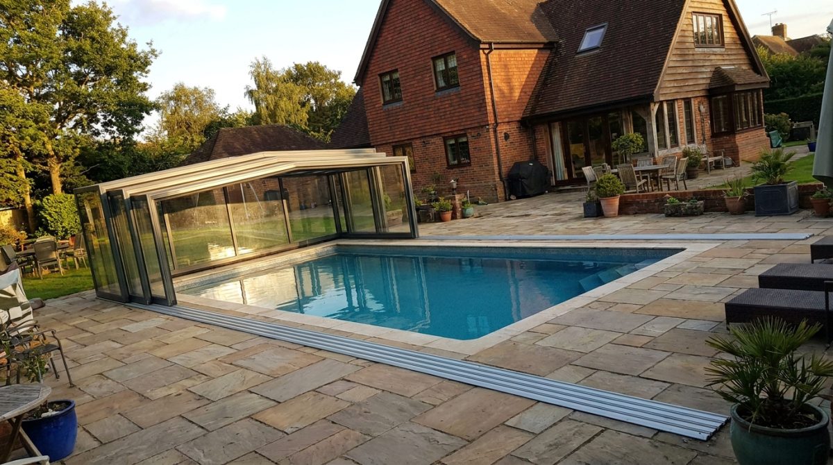 A well-lit composite swimming pool with glass enclosure partially open, surrounded by stone paving on a UK residential property, photographed in late afternoon light